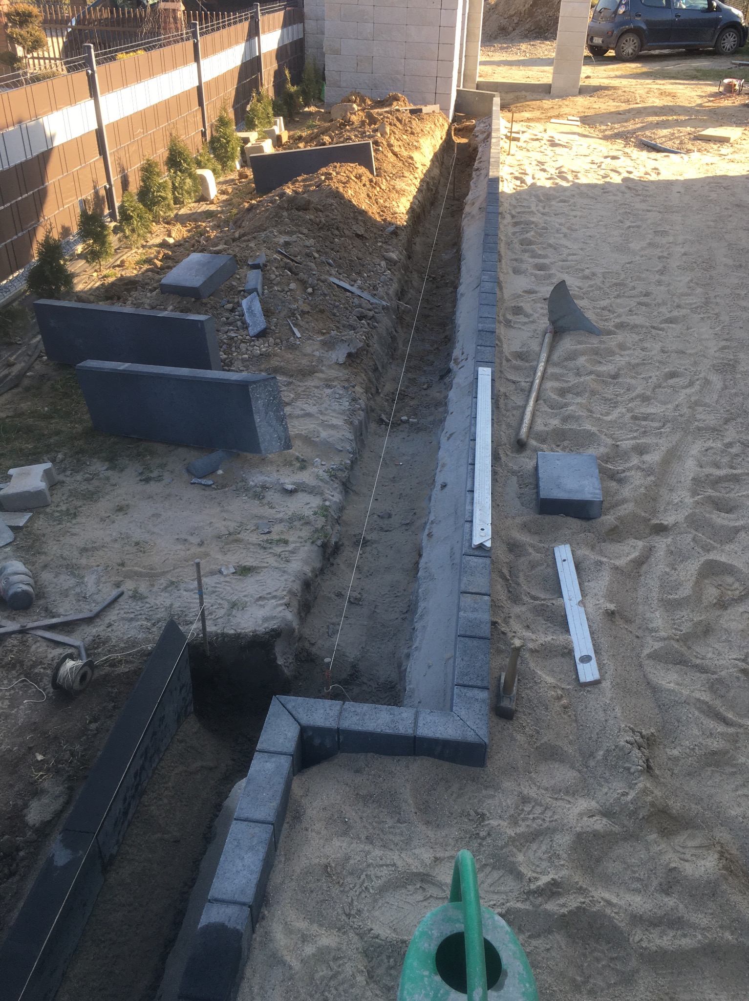 Przygotowany wykop pod obrzeża z ułożonymi elementami z kostki granitowej, widoczne narzędzia, sznurek wyznaczający linię, poziomica i zielona konewka na piaszczystym podłożu.