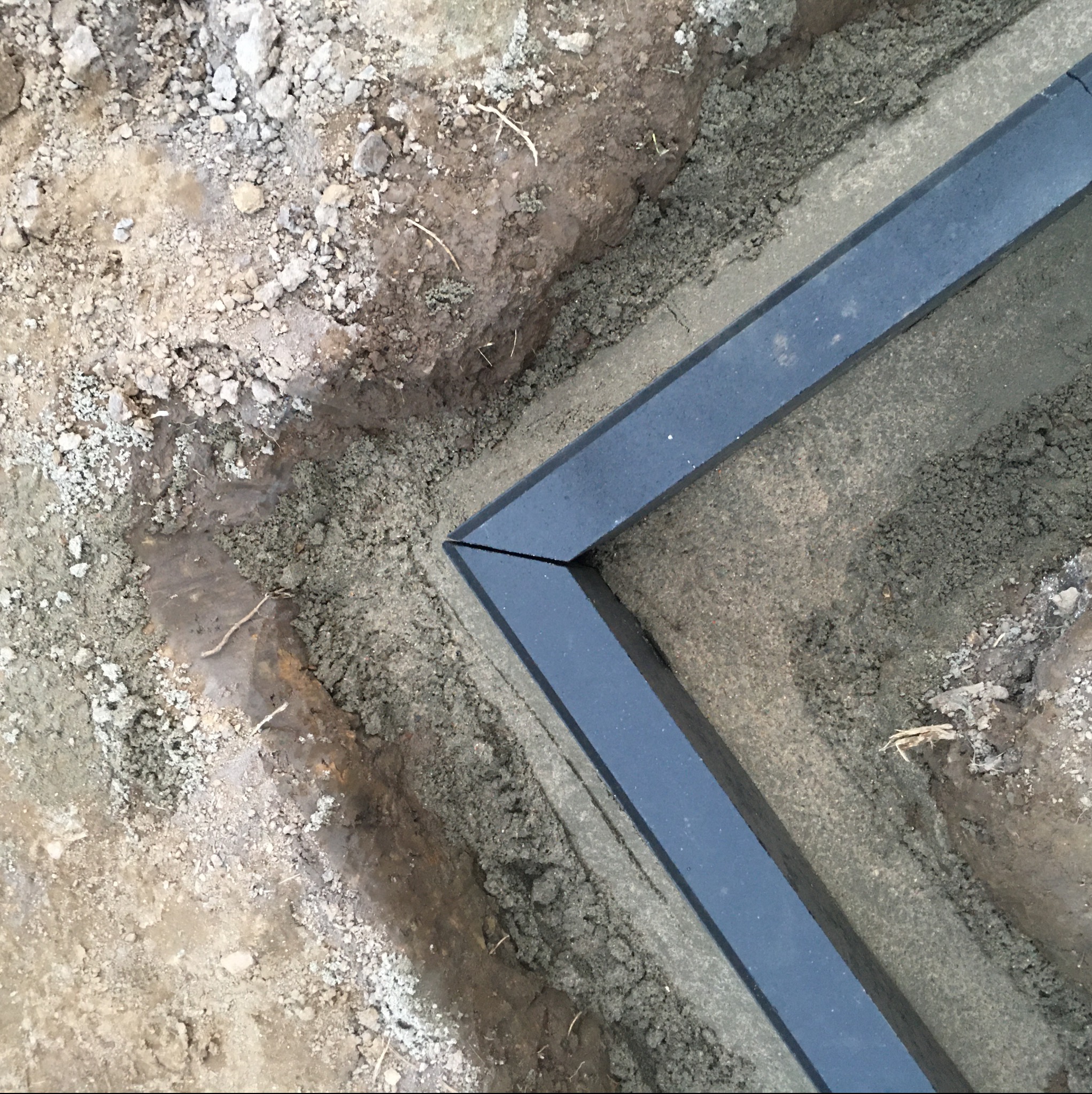 Fragment wykopu z ułożonym obrzeżem z ciemnoszarej kostki betonowej, widoczne nierówności terenu i przygotowanie podłoża.