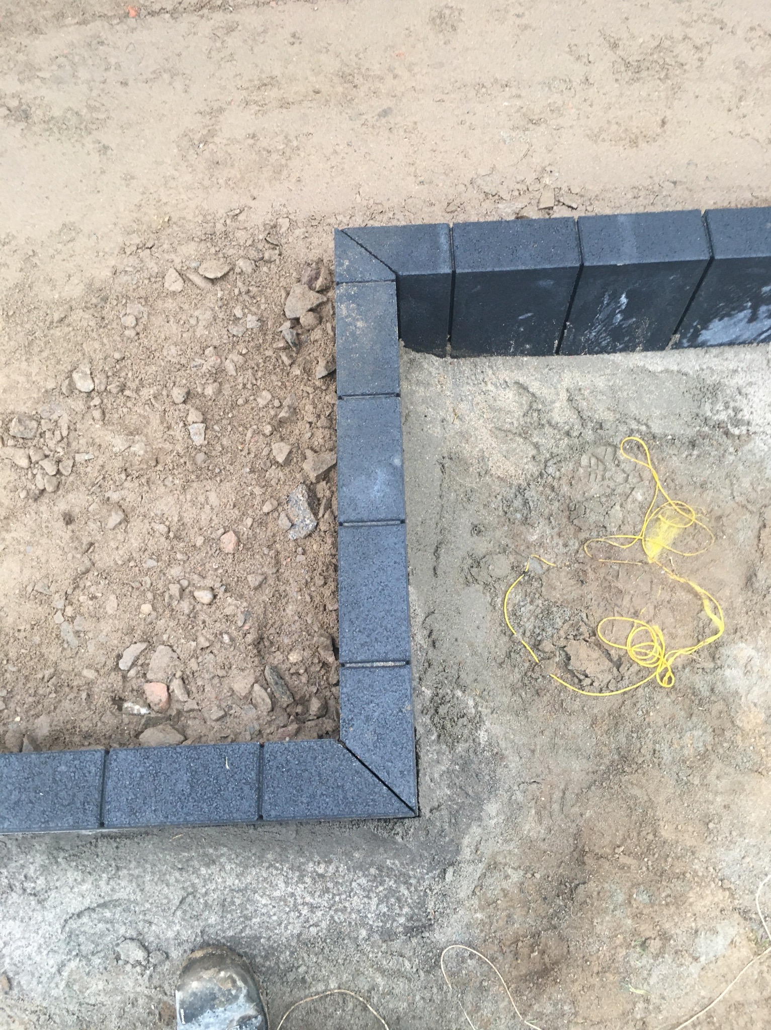 Krawężnik z ciemnej kostki obramowujący obszar przygotowany pod nawierzchnię, widoczny fragment buta roboczego i żółty sznurek wyznaczający linię.