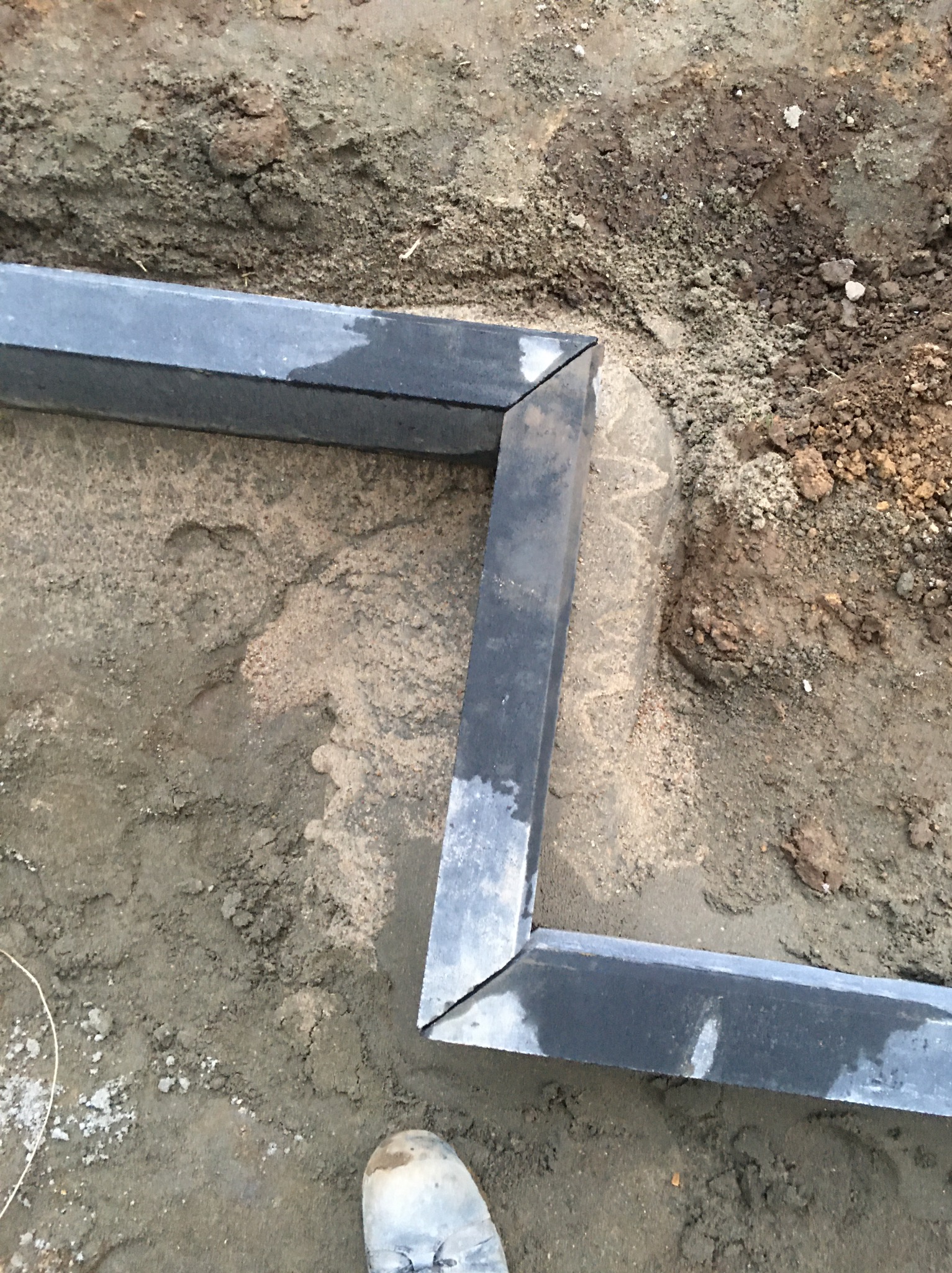 Fragment obramowania z ciemnych, mokrych elementów ułożonych w kształt litery 'Z' na piasku i ziemi w trakcie prac ziemnych, widoczny fragment roboczego buta.