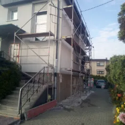 Dom jednorodzinny w trakcie prac elewacyjnych, widoczne rusztowania, świeżo pomalowana szara elewacja z elementami dekoracyjnymi na cokole i pod dachem, kostka brukowa na podjeździe.