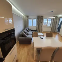 Jasne wnętrze mieszkania z parkietem, szarą sofą narożną, białym stołem i krzesłami z tapicerowanymi oparciami, widok na okna z zasłonami i zabudowaną kuchnią z piekarnikiem.