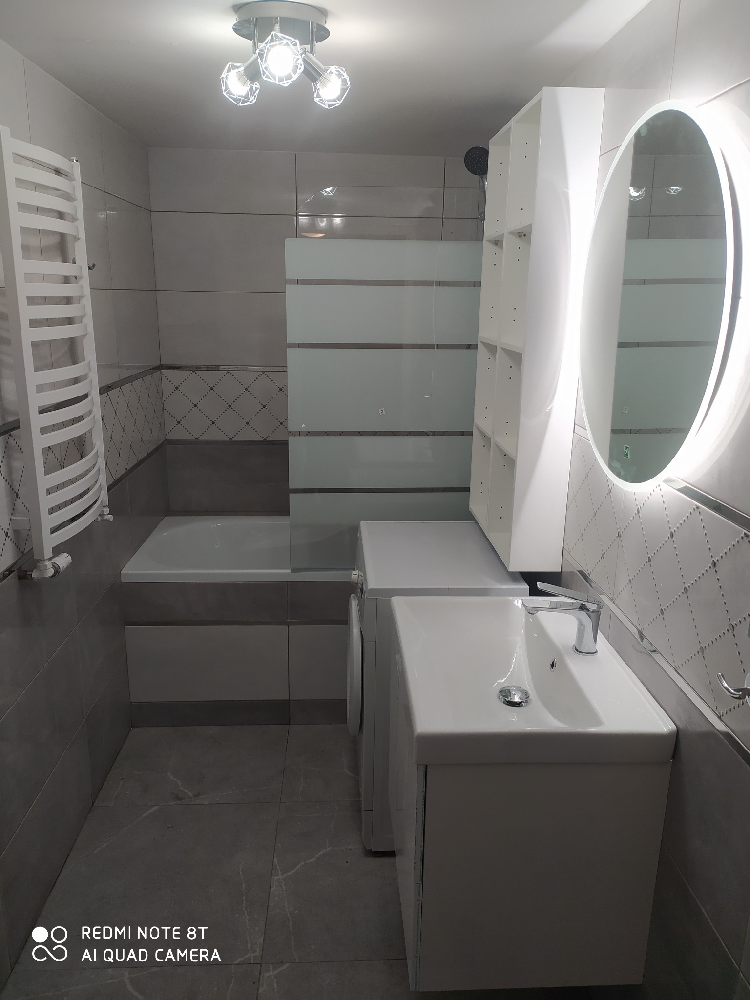 Nowoczesna łazienka z owalnym lustrem z podświetleniem LED, białą umywalką z szafką, grzejnikiem drabinkowym i płytkami w odcieniach szarości z dekoracyjnym wzorem.