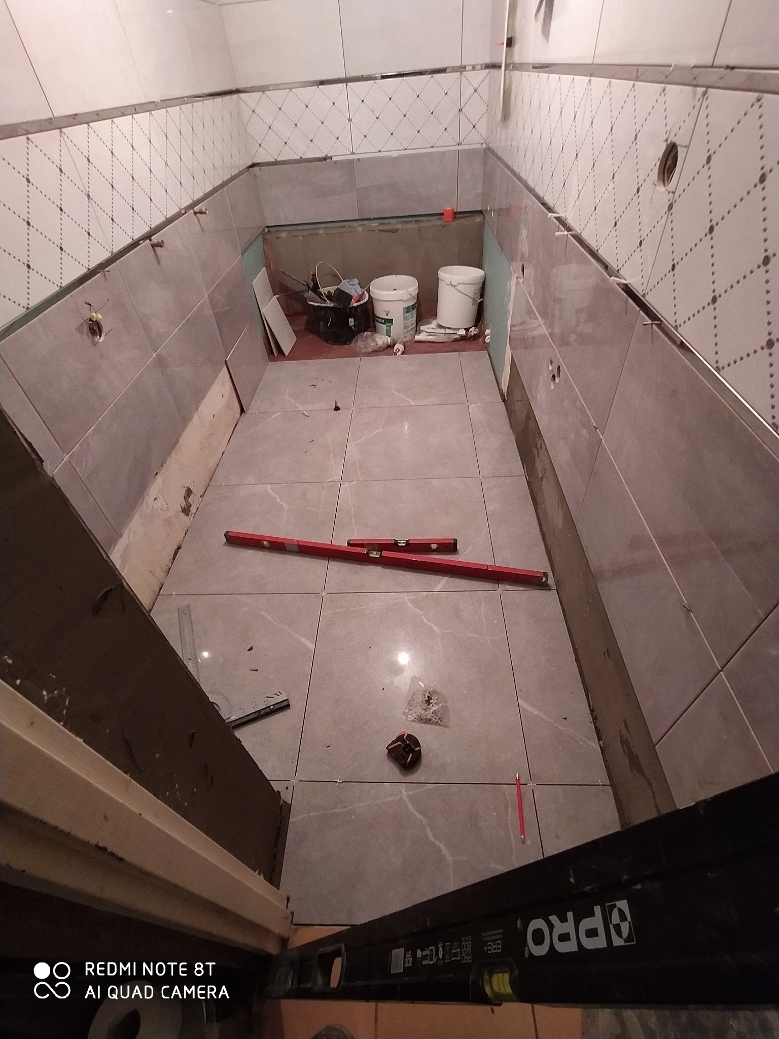 Łazienka w trakcie prac: położone szare płytki podłogowe, poziomica, ściany częściowo wyłożone płytkami z dekoracyjnym paskiem, widoczne narzędzia i materiały budowlane.