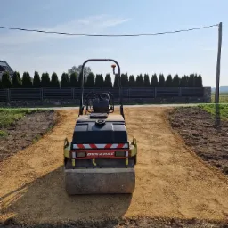 Kompaktor wibracyjny Dynapac na przygotowanym podłożu z kruszywa, widoczny na tle ogrodzenia z tujami i słupem energetycznym.