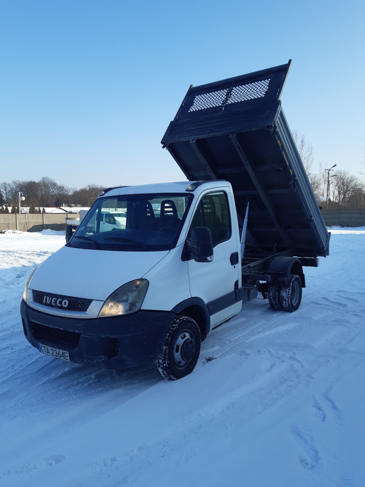 Biały samochód ciężarowy Iveco z otwartą, pustą skrzynią ładunkową, stojący na śniegu w słoneczny dzień.