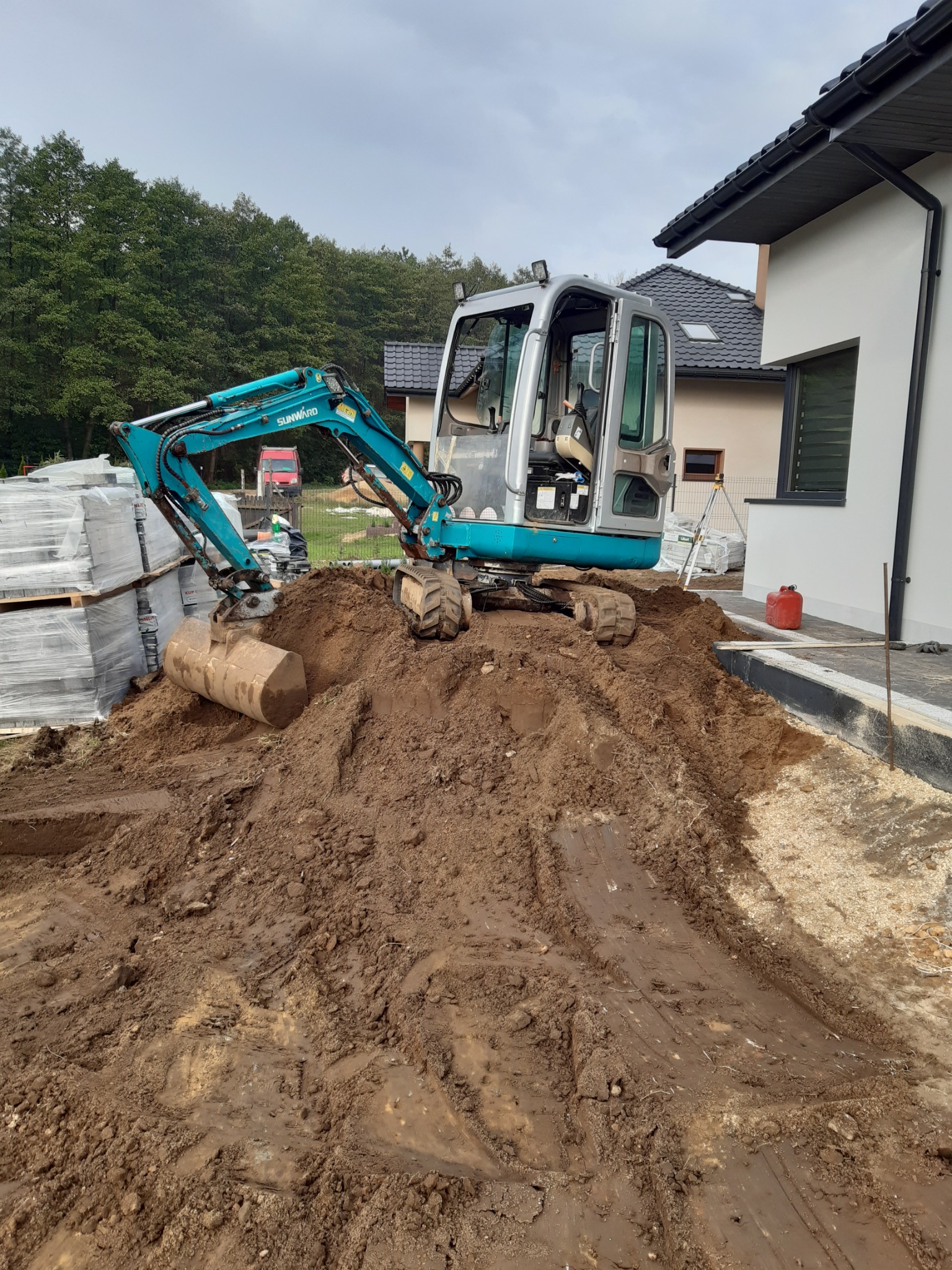 Mała, turkusowa koparka Sunward stoi na stercie ziemi obok nowego domu w trakcie budowy. W tle zaparkowany czerwony dostawczy samochód i palety z materiałami budowlanymi.