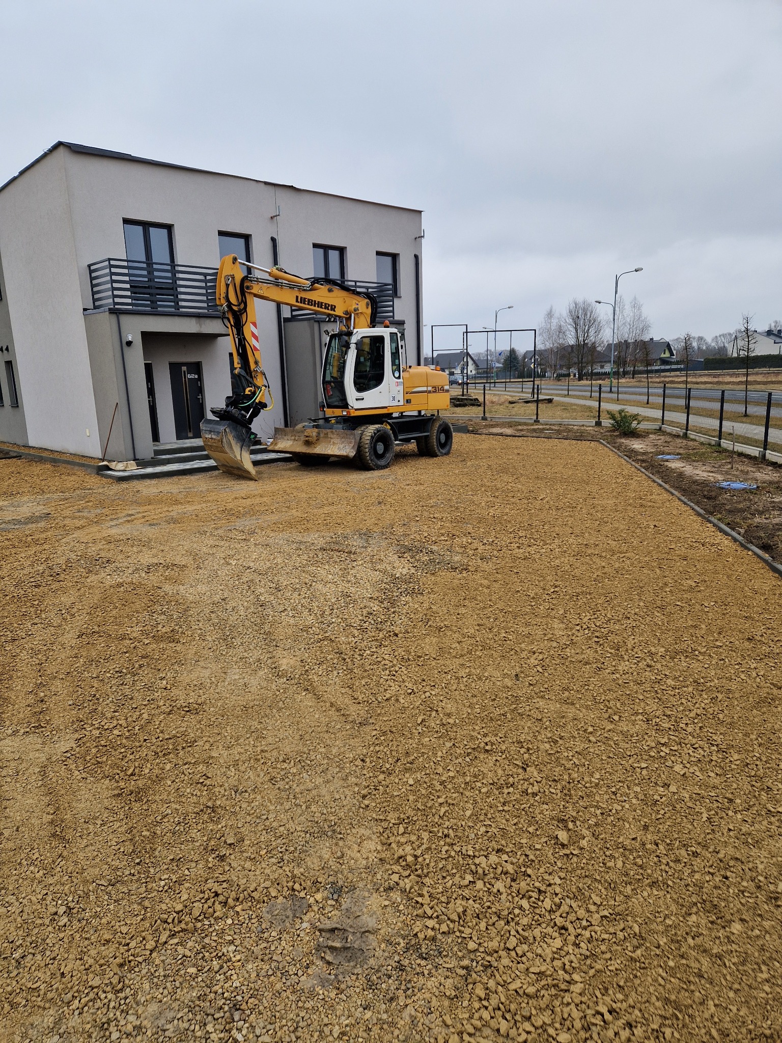 Żółta koparka Liebherr na placu budowy przed nowoczesnym domem, teren wyrównany drobnym kruszywem, widoczne ślady opon i świeżo ułożone obrzeża.