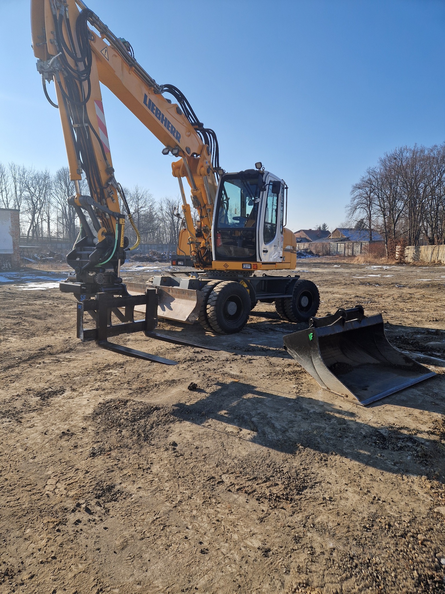 Żółto-biała koparka Liebherr z zamontowanymi widłami i łyżką na ziemnym placu budowy w słoneczny dzień.