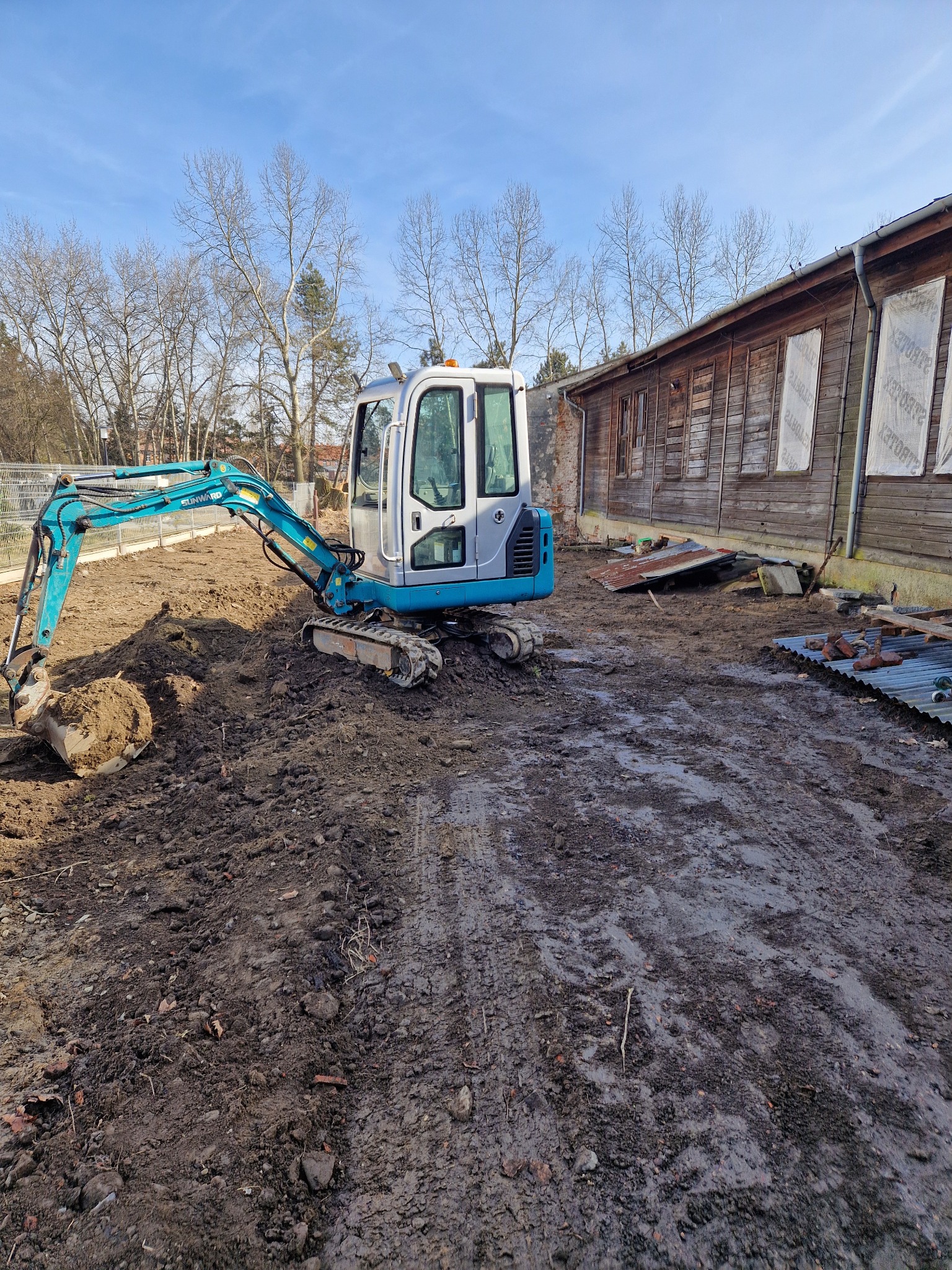 Mała, niebiesko-biała koparka na gąsienicach, pracująca na placu budowy z brudną ziemią i starym drewnianym budynkiem w tle, w słoneczny dzień.
