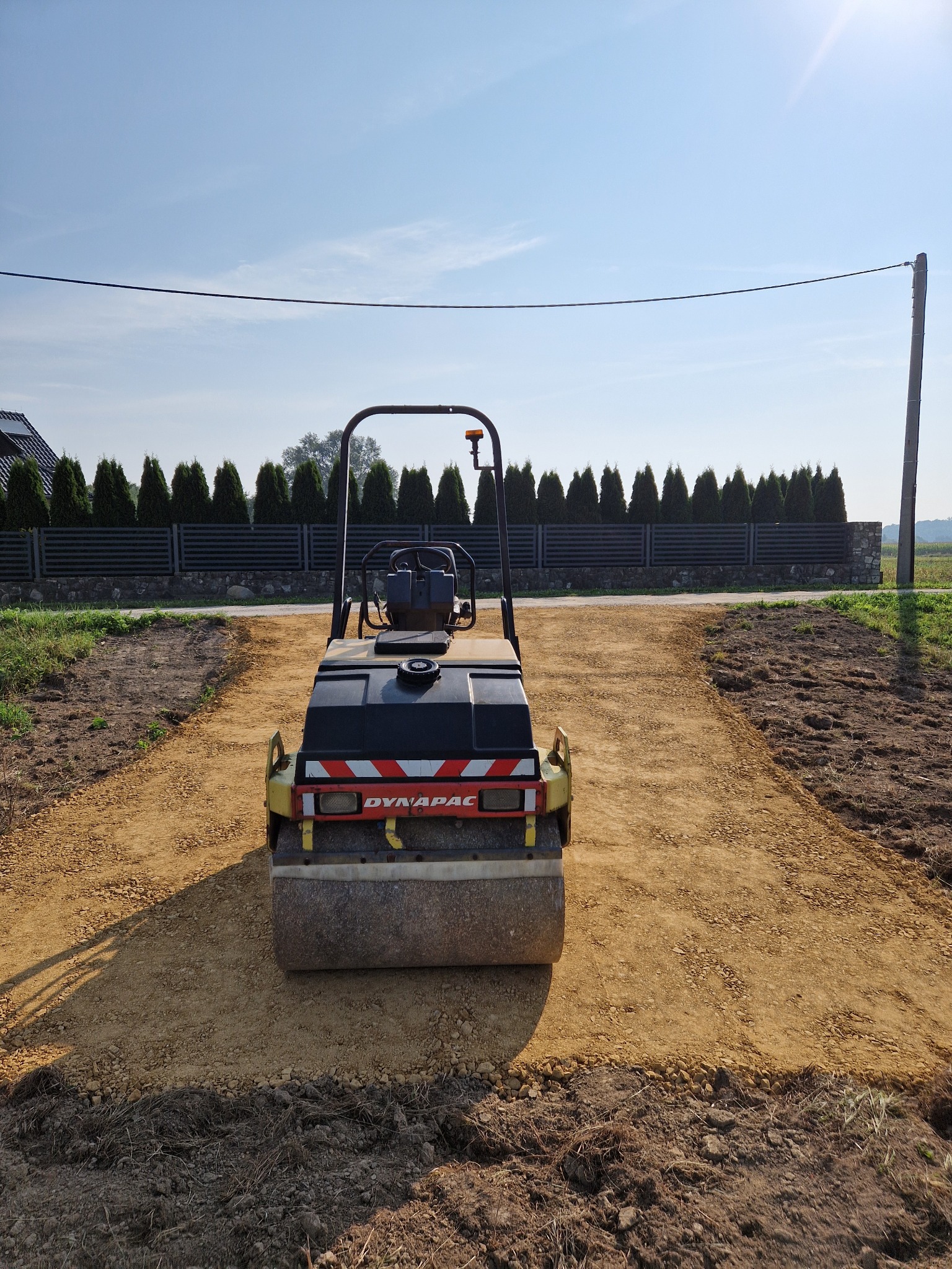 Kompaktor wibracyjny Dynapac na przygotowanym podłożu z kruszywa, widoczny na tle ogrodzenia z tujami i słupem energetycznym.