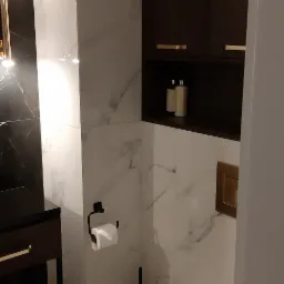 Nowoczesna łazienka z białymi marmurkowymi płytkami, wiszącą toaletą, czarną szczotką do WC i szafką z ciemnego drewna z złotymi uchwytami.