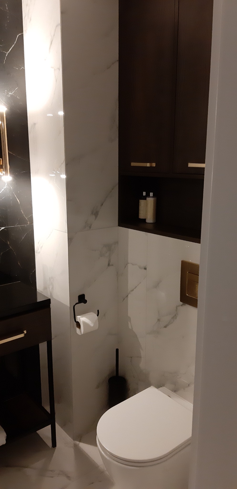 Nowoczesna łazienka z białymi marmurkowymi płytkami, wiszącą toaletą, czarną szczotką do WC i szafką z ciemnego drewna z złotymi uchwytami.