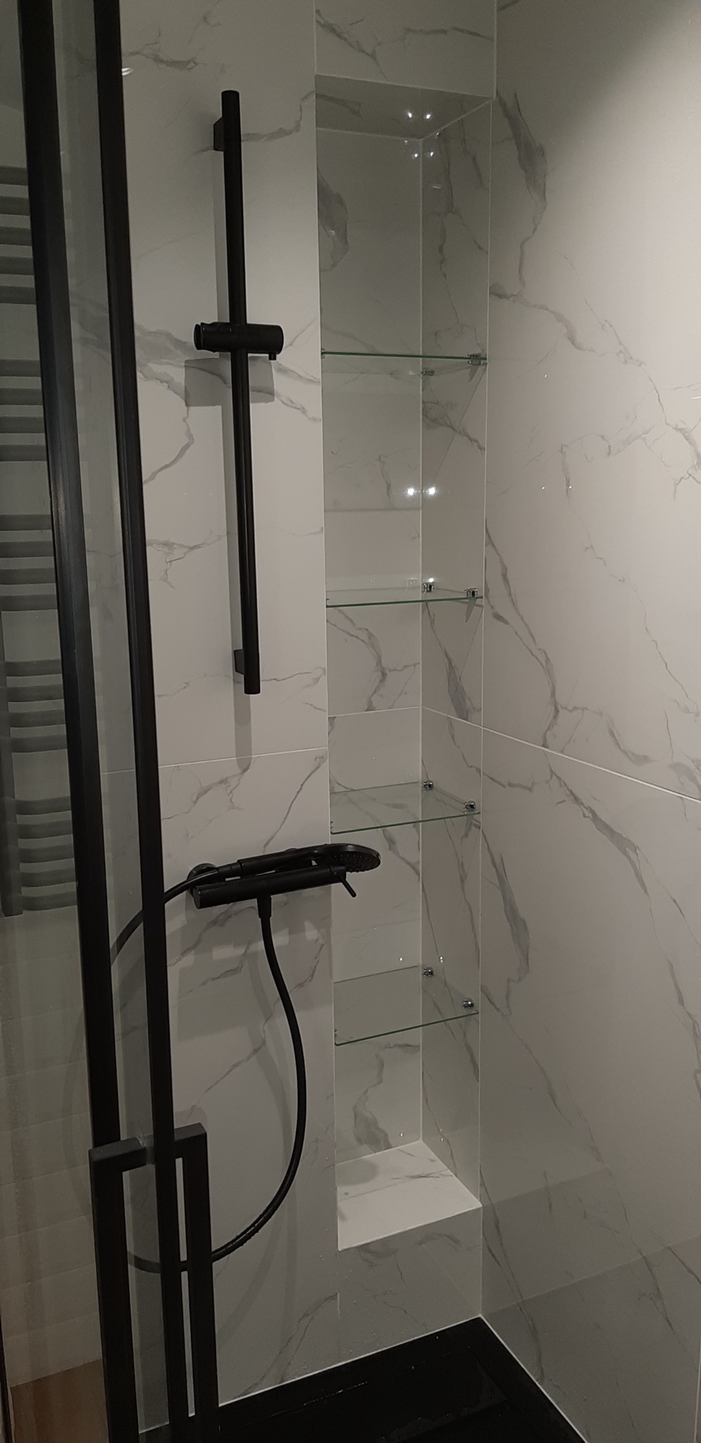 Nowoczesna łazienka z kabiną prysznicową z czarnymi elementami, białe płytki imitujące marmur, wnęka z szklanymi półkami.