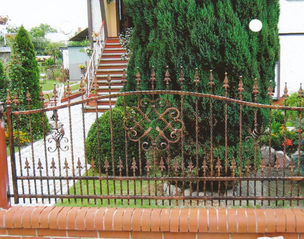 Kute ogrodzenie w kolorze miedzi z ozdobnymi elementami na tle zielonego ogrodu z widocznymi schodami prowadzącymi do domu.