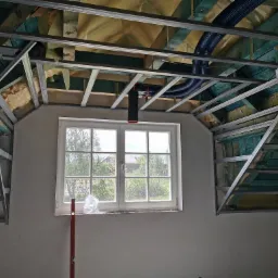 Stelaż z profili metalowych pod zabudowę poddasza, widoczna izolacja, niebieskie rury wentylacyjne i okno z widokiem na zieleń.
