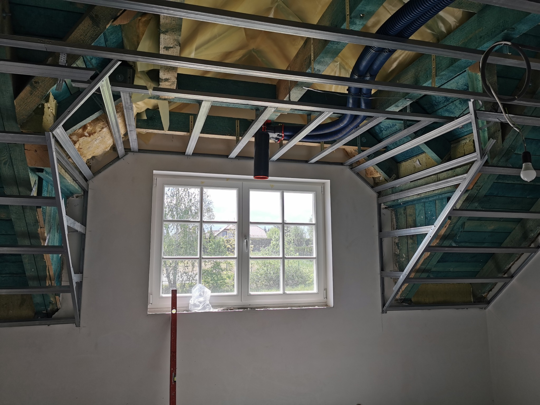 Stelaż z profili metalowych pod zabudowę poddasza, widoczna izolacja, niebieskie rury wentylacyjne i okno z widokiem na zieleń.