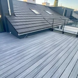 Szary taras na dachu budynku z metalową balustradą, widok na dachy sąsiednich budynków w słoneczny dzień.