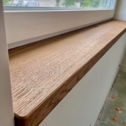 PURITY ADAM STACHURA - Zamontowany drewniany parapet wewnętrzny o zaokrąglonych krawędziach, widoczny z bliska, z oknem w tle. Jasne, naturalne oświetlenie podkreśla strukturę drewna.