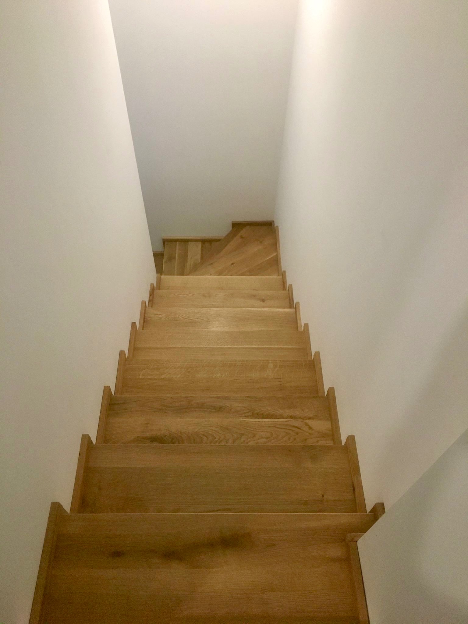 Jasne, drewniane schody z minimalistycznymi stopniami, prowadzące w górę w białym wnętrzu. Widok z góry, podkreślający naturalną teksturę drewna i prostotę konstrukcji.