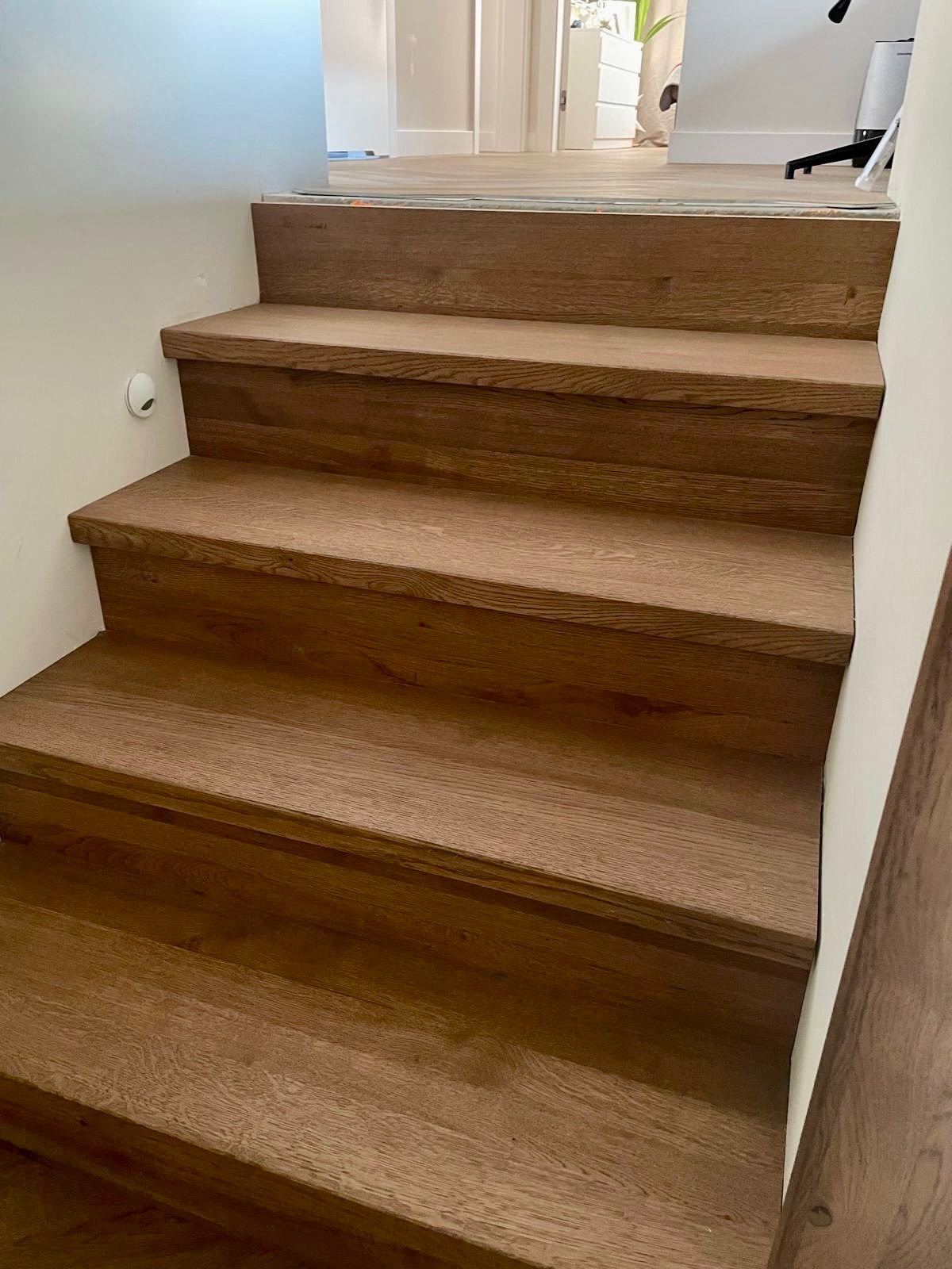 Jasne, drewniane schody wewnętrzne z widocznym usłojeniem drewna, prowadzące do góry. Minimalistyczny design, białe ściany, ciepłe oświetlenie.