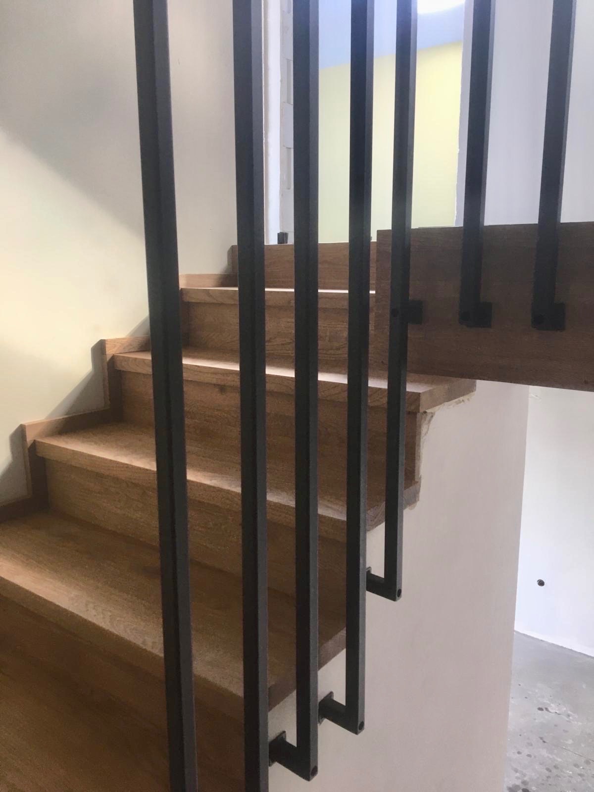Drewniane schody z nowoczesną, czarną balustradą. Stopnie z jasnego drewna, pionowe elementy balustrady o kwadratowym przekroju. Minimalistyczny design.