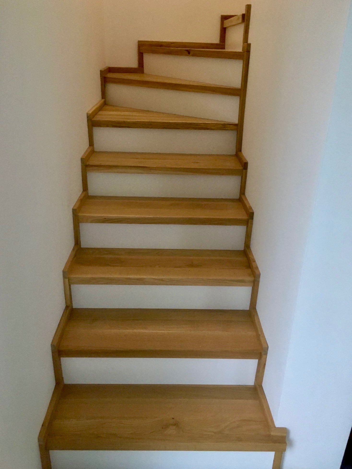 Drewniane schody z jasnego drewna, widok z dołu, z białymi ścianami po bokach. Stopnie schodów są wyraźnie widoczne, a konstrukcja wydaje się być solidna i estetyczna.