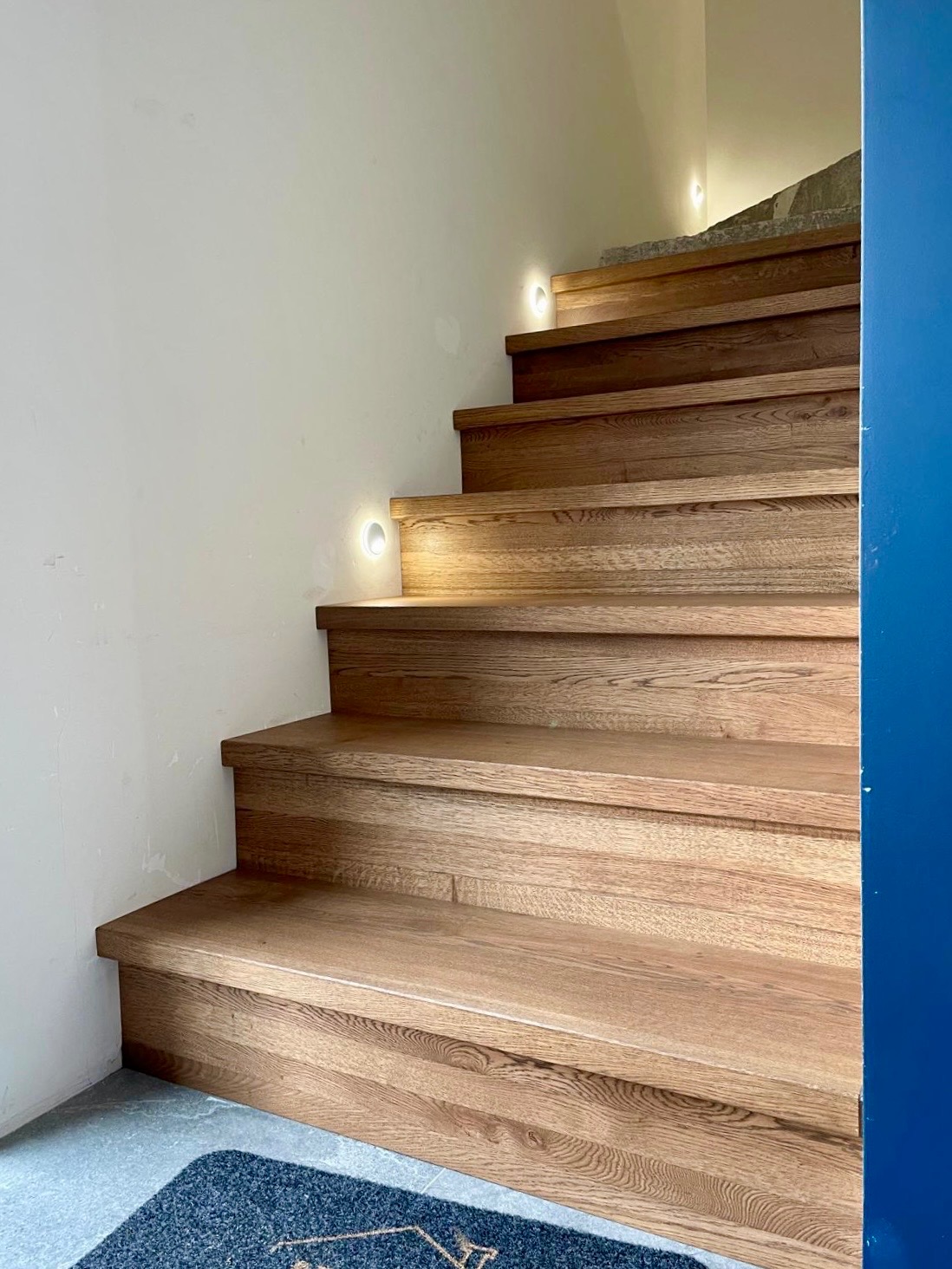 Dębowe schody z podświetleniem LED wzdłuż białej ściany, widoczne z dołu, z fragmentem niebieskiej ściany po prawej stronie i wycieraczką u dołu.