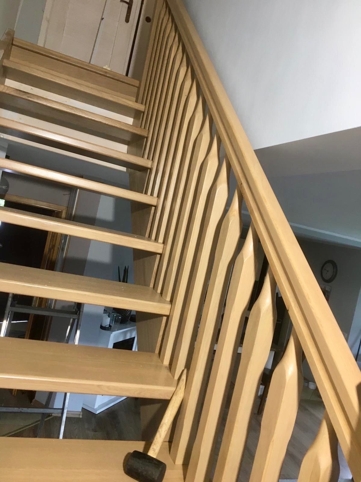 Jasne, drewniane schody z ozdobną balustradą, widziane z dołu. Na jednym ze stopni leży czarny młotek. Widoczne elementy wystroju wnętrza w tle.