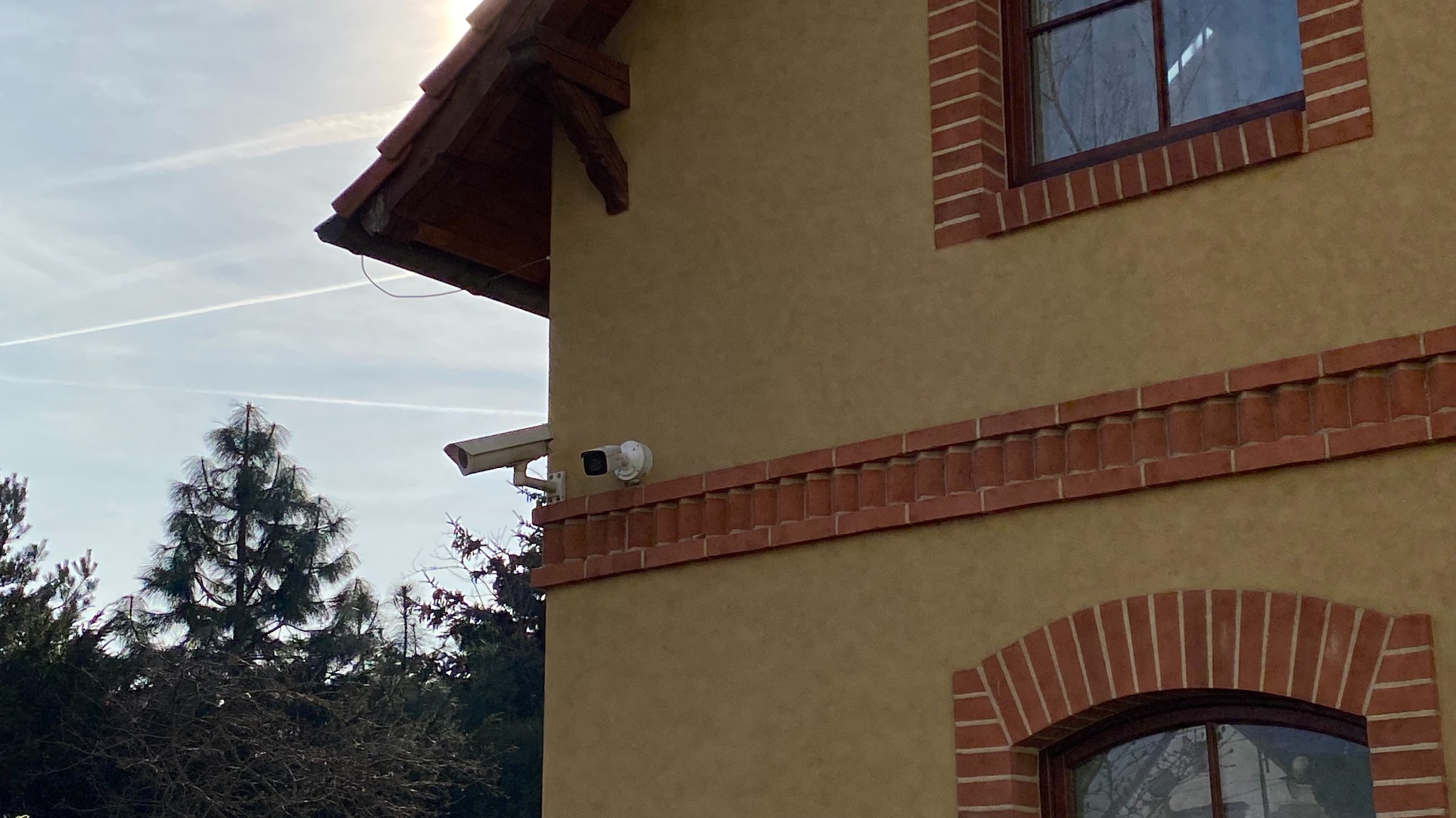 Dwie kamery monitoringu zamontowane na elewacji budynku z beżowym tynkiem i ceglanymi elementami dekoracyjnymi, widoczne drzewa i niebo z smugami kondensacyjnymi.