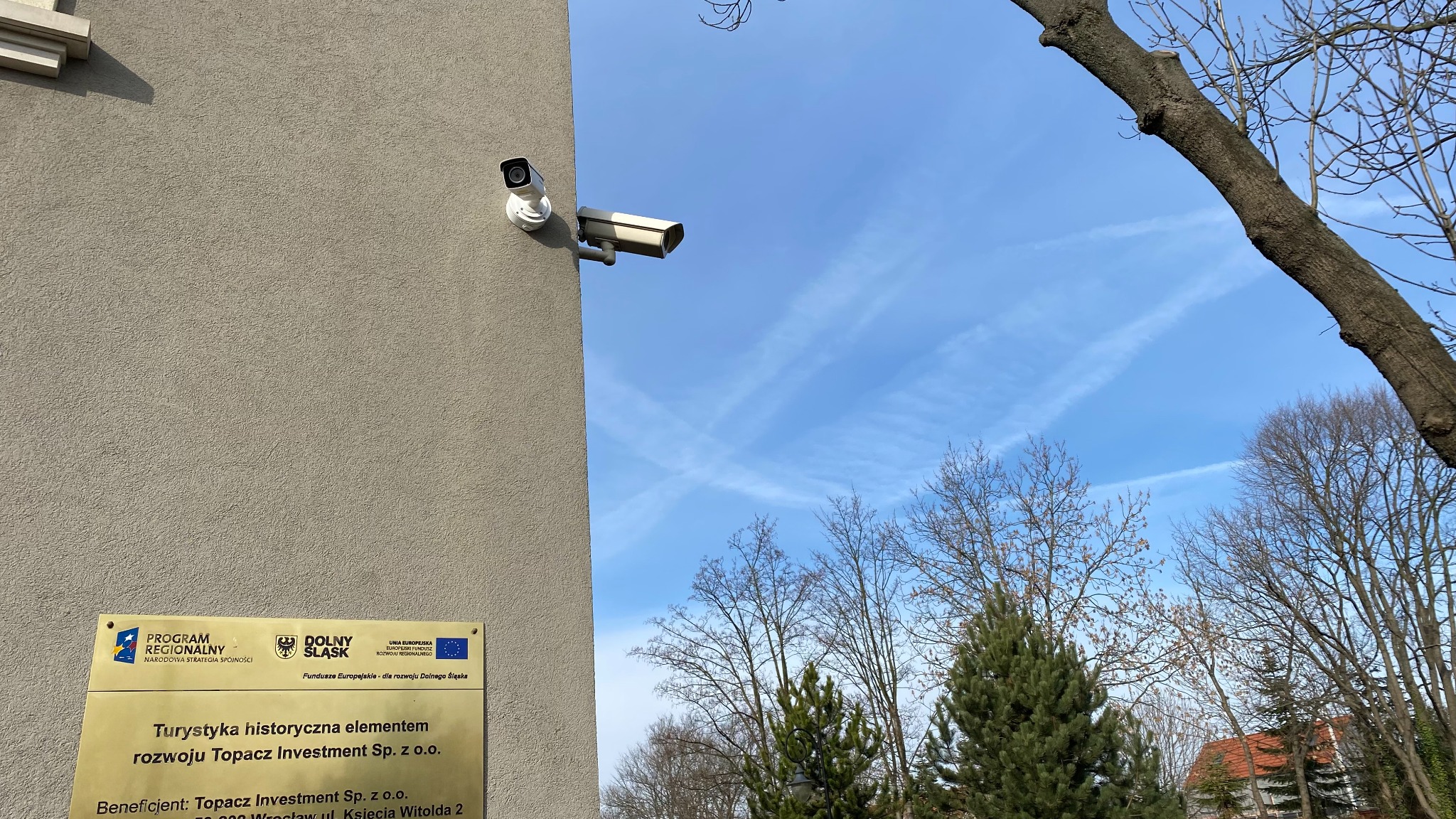 Kamera monitoringu przymocowana do ściany budynku, skierowana w stronę błękitnego nieba z widocznymi smugami kondensacyjnymi samolotów, obok tablica informacyjna z logo Dolnego Śląska.
