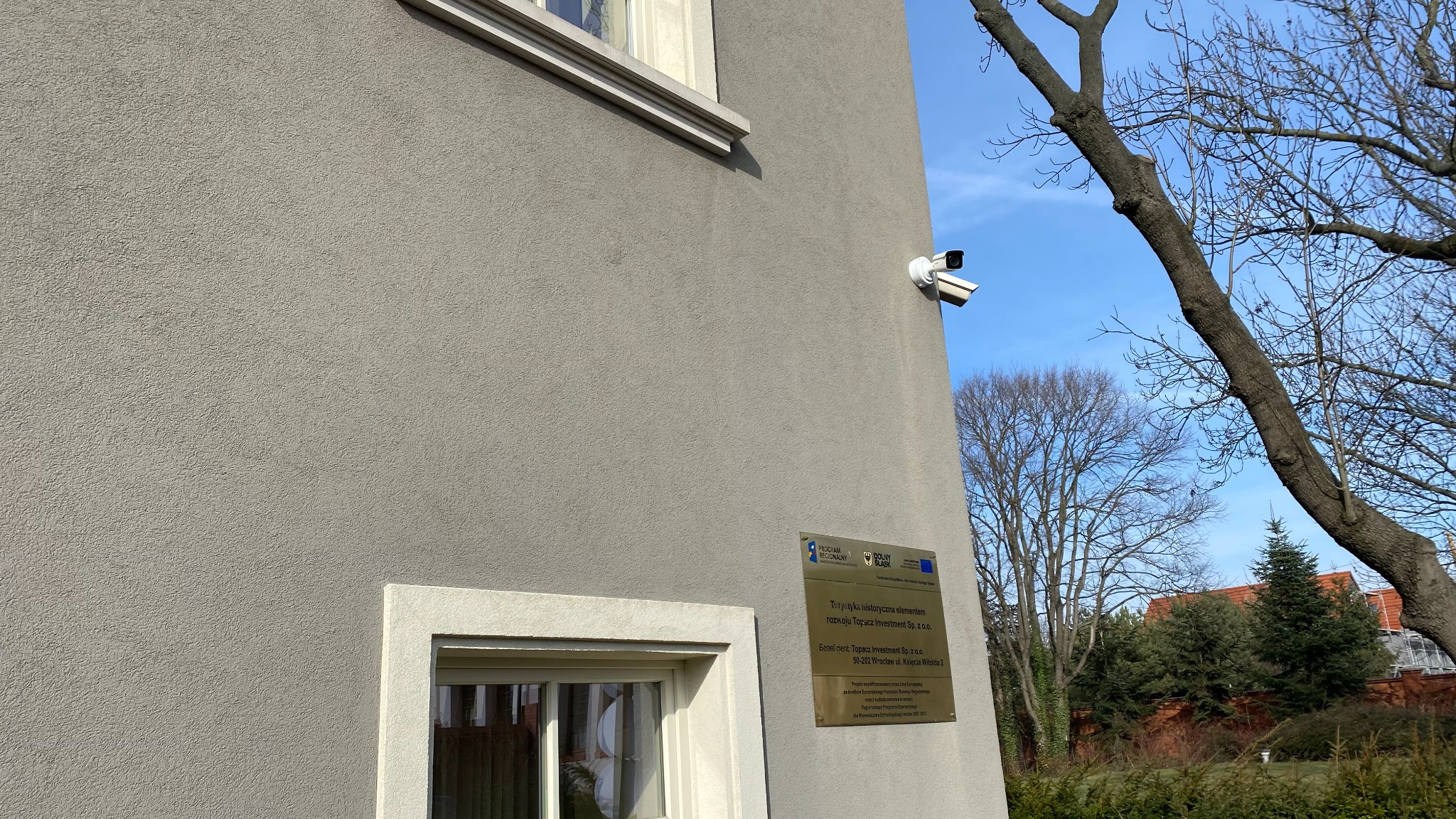 Dwie kamery monitoringu zamontowane na szarej ścianie budynku obok okna i tabliczki informacyjnej, widok z dołu na tle błękitnego nieba i gałęzi drzew.