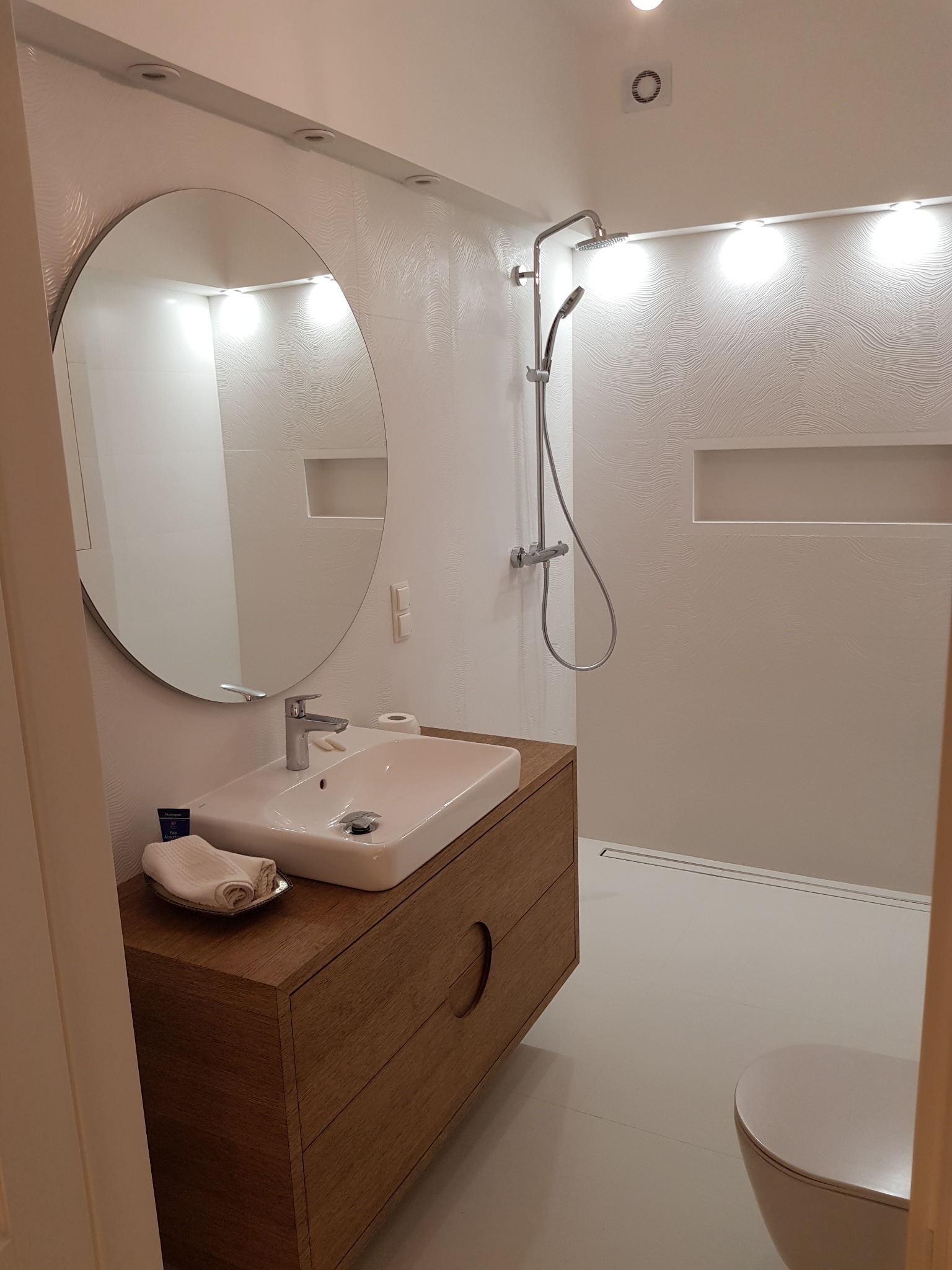 Nowoczesna łazienka z umywalką nablatową na drewnianej szafce, okrągłym lustrem i kabiną prysznicową typu walk-in z odpływem liniowym, oświetlona punktowymi lampami sufitowymi.