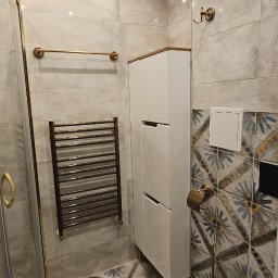 RADOSŁAW SZYNIEC - Ukończona łazienka z kabiną prysznicową ze złotymi detalami, grzejnikiem drabinkowym w stylu retro, białą szafką i wzorzystymi płytkami na podłodze i części ścian.