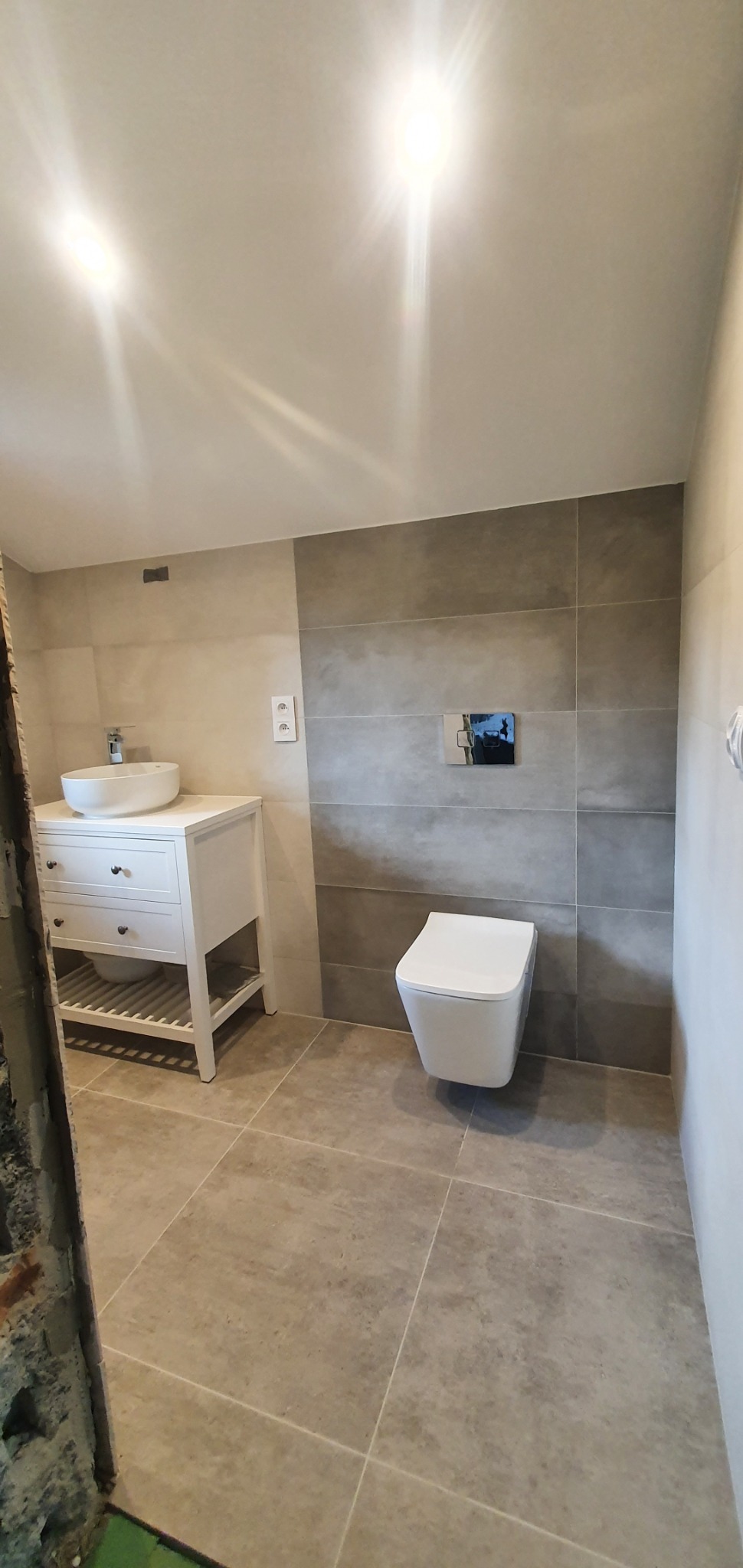 Nowoczesna łazienka z szarymi płytkami, wiszącą toaletą i białą szafką z umywalką, widok na fragment niedokończonego wejścia.