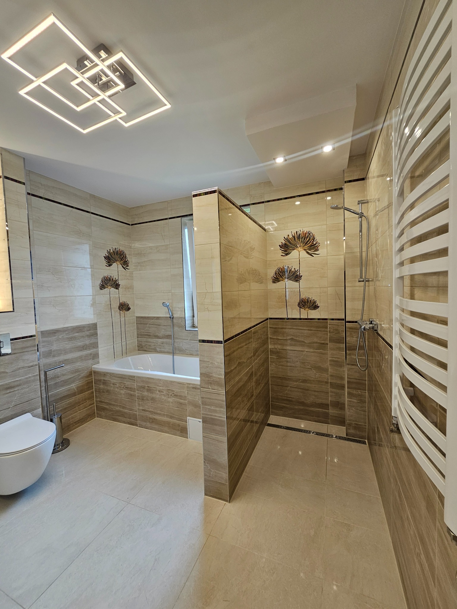 Wykończona łazienka z beżowymi płytkami imitującymi drewno, kabiną prysznicową typu walk-in, wanną, wiszącą toaletą i grzejnikiem drabinkowym; dekoracyjne motywy kwiatowe na płytkach.
