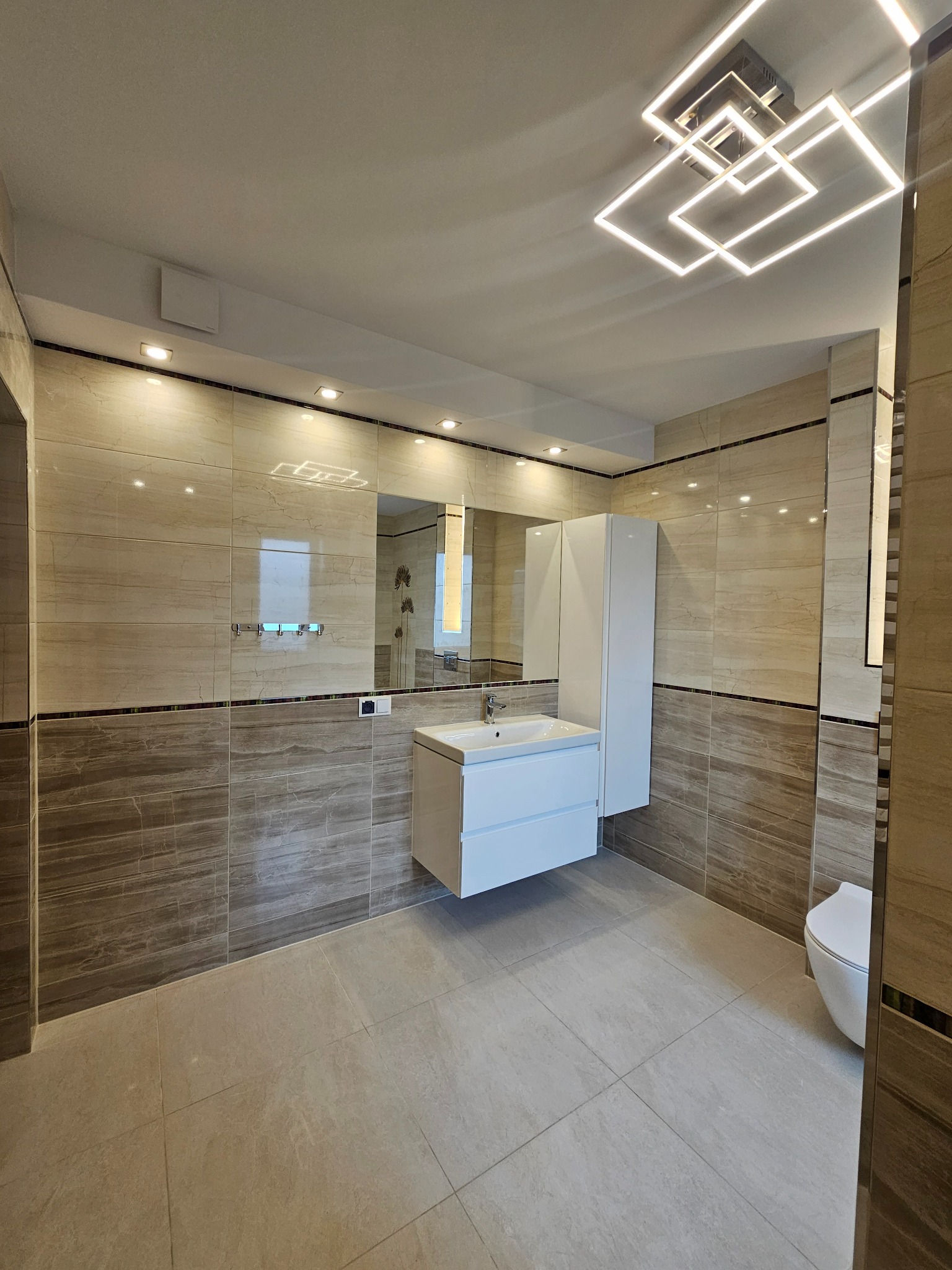 Wyremontowana łazienka z beżowo-brązowymi płytkami imitującymi drewno, białą szafką umywalkową i geometryczną lampą sufitową LED.
