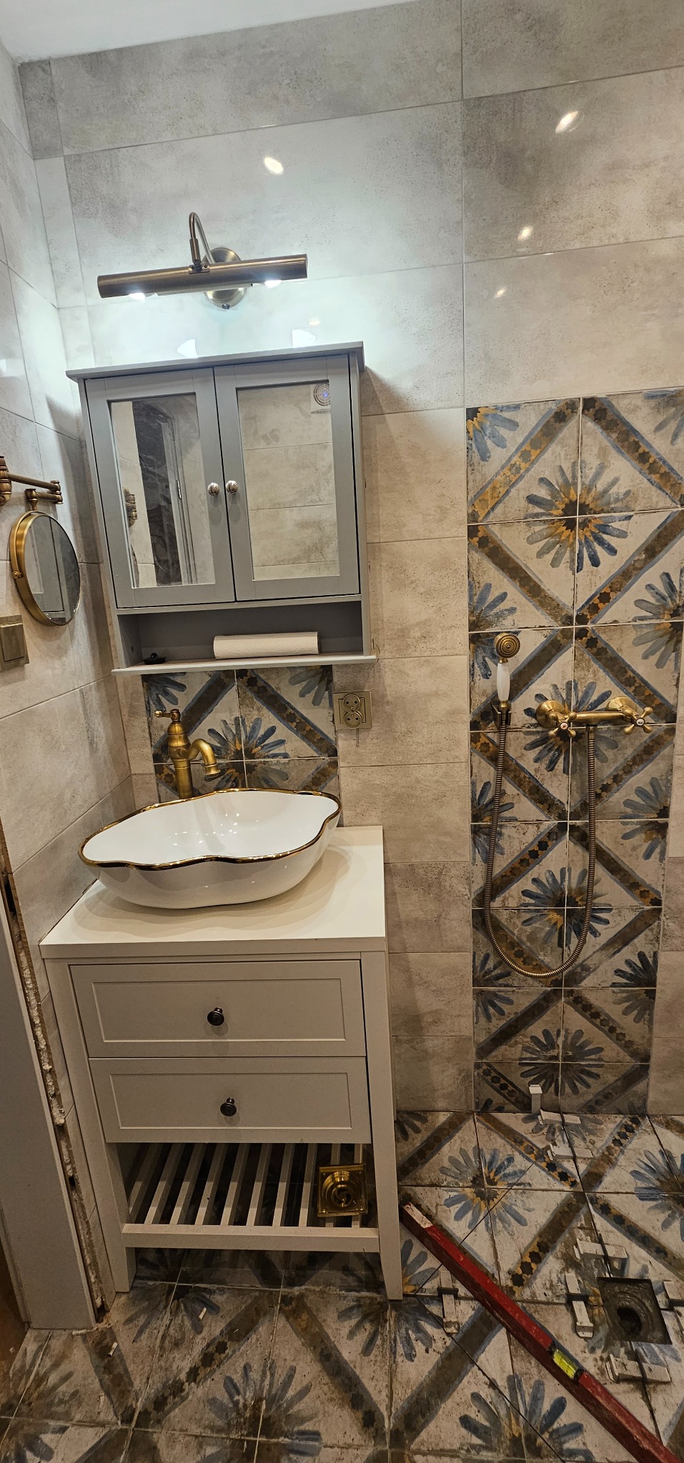 Łazienka w trakcie renowacji: biała umywalka z dekoracyjnym złotym obramowaniem na szafce z szufladami, złota bateria, lustro w szarej szafce, ozdobne płytki na ścianie i podłodze, widoczne ślady...