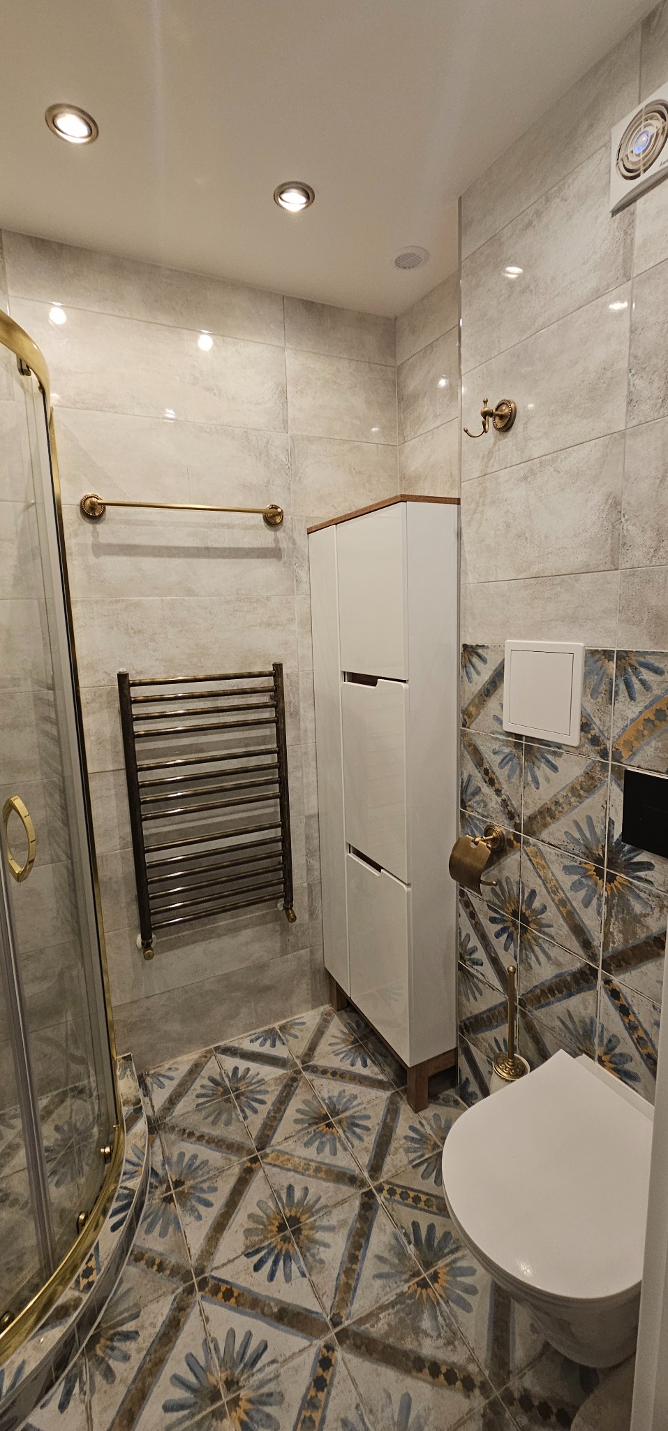 Ukończona łazienka z kabiną prysznicową ze złotymi detalami, grzejnikiem drabinkowym w stylu retro, białą szafką i wzorzystymi płytkami na podłodze i części ścian.