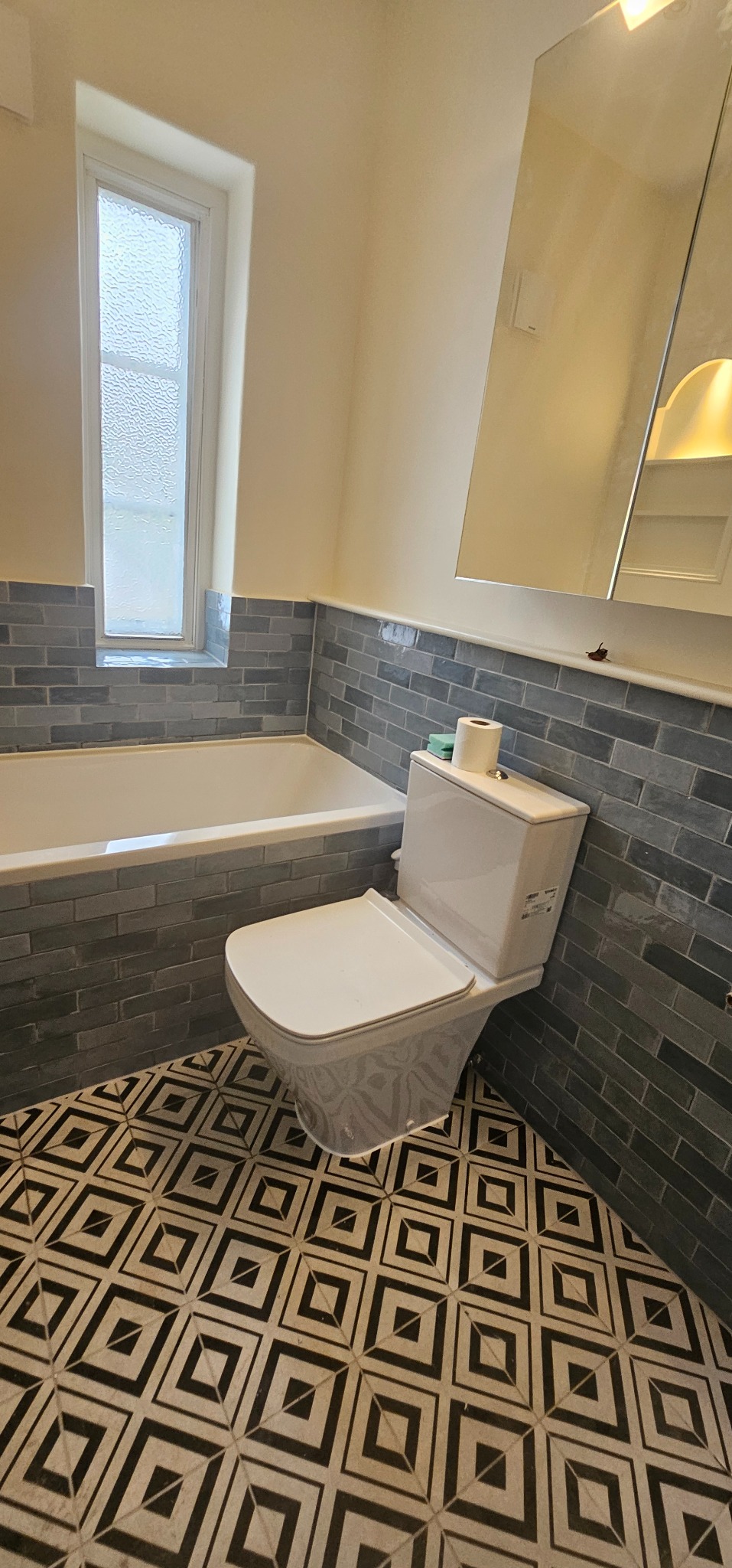Wykończona łazienka z szaro-niebieską cegłą nad wanną i toaletą, oraz wzorzystą czarno-białą podłogą, widok na okno z mleczną szybą i lustro z oświetleniem.