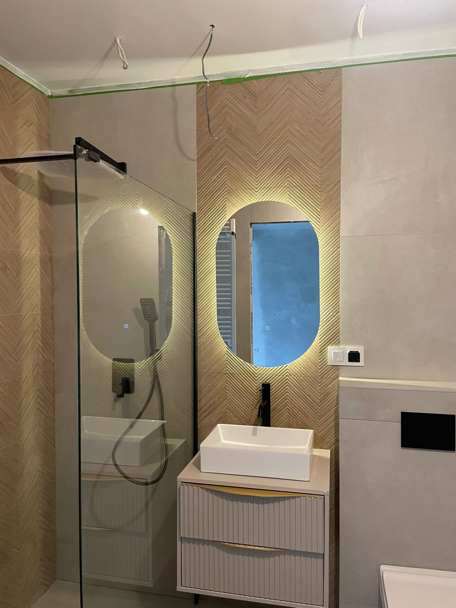 Nowoczesna łazienka z podświetlanym owalnym lustrem, wzorzystą ścianą w jodełkę, białą umywalką nablatową i szklaną kabiną prysznicową. Widoczne niedokończone prace elektryczne na suficie.