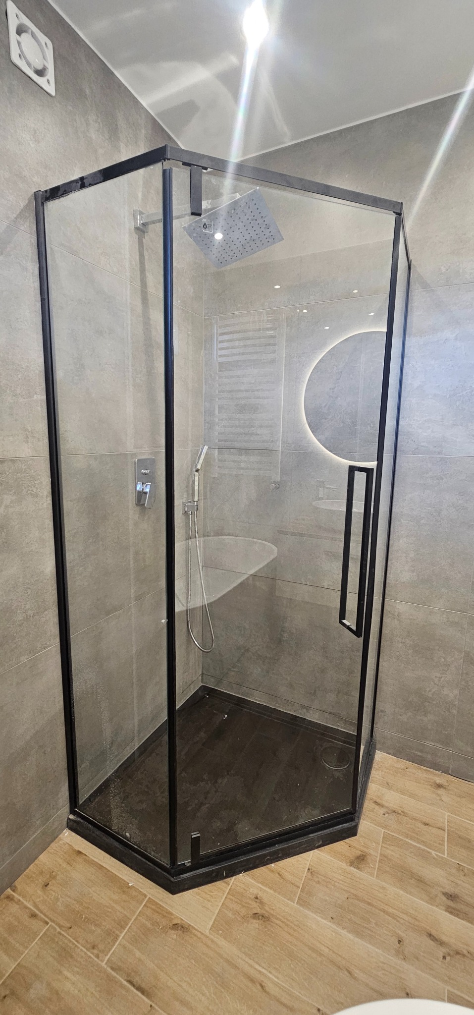 Nowoczesna kabina prysznicowa z czarnymi profilami, deszczownicą i słuchawką, szare płytki na ścianach, podświetlane okrągłe lustro w tle, drewnopodobna podłoga w jodełkę.