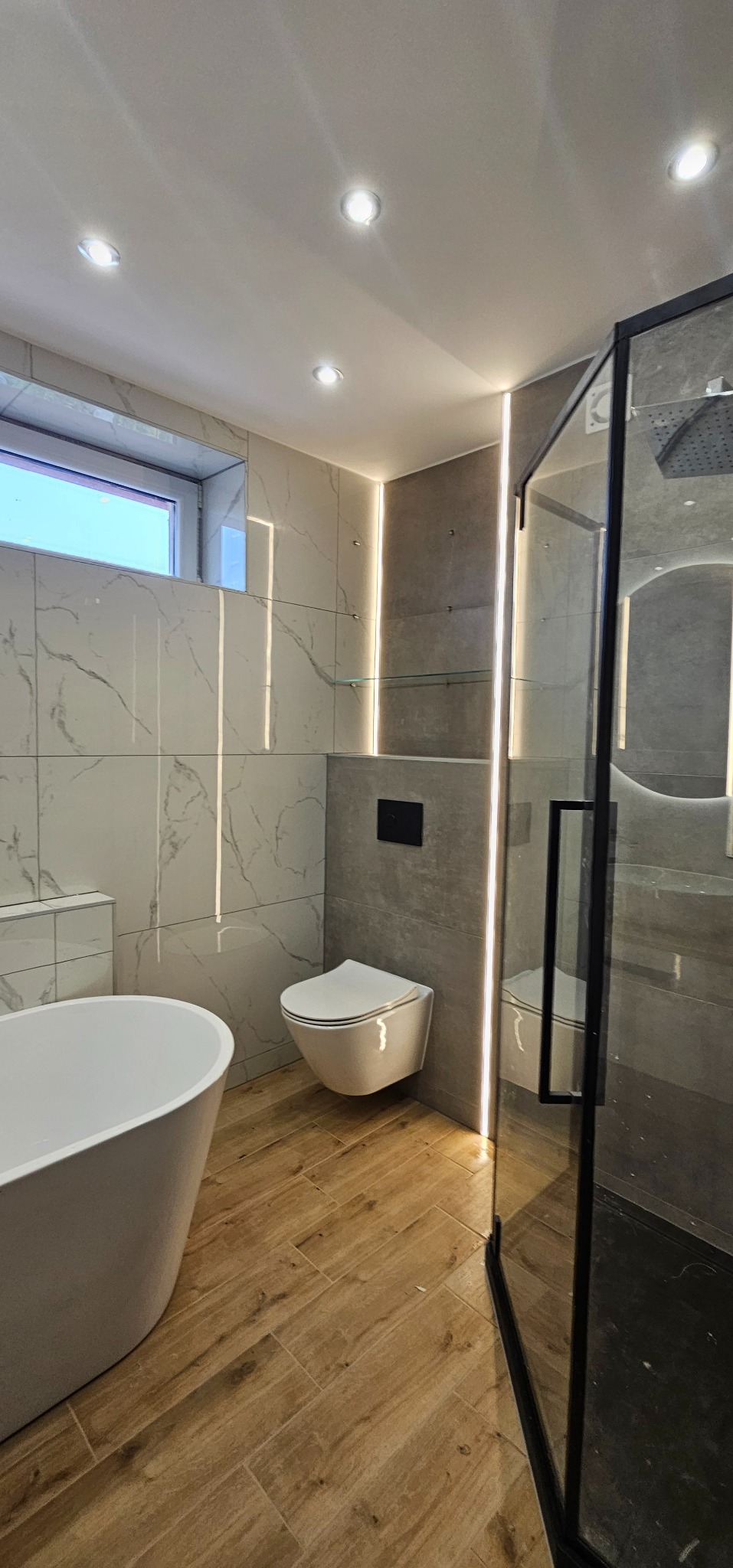 Nowoczesna łazienka z wolnostojącą wanną, podwieszaną toaletą i kabiną prysznicową z czarnymi ramami, oświetlona listwami LED i punktowymi lampami sufitowymi, wykończona płytkami imitującymi marmur...