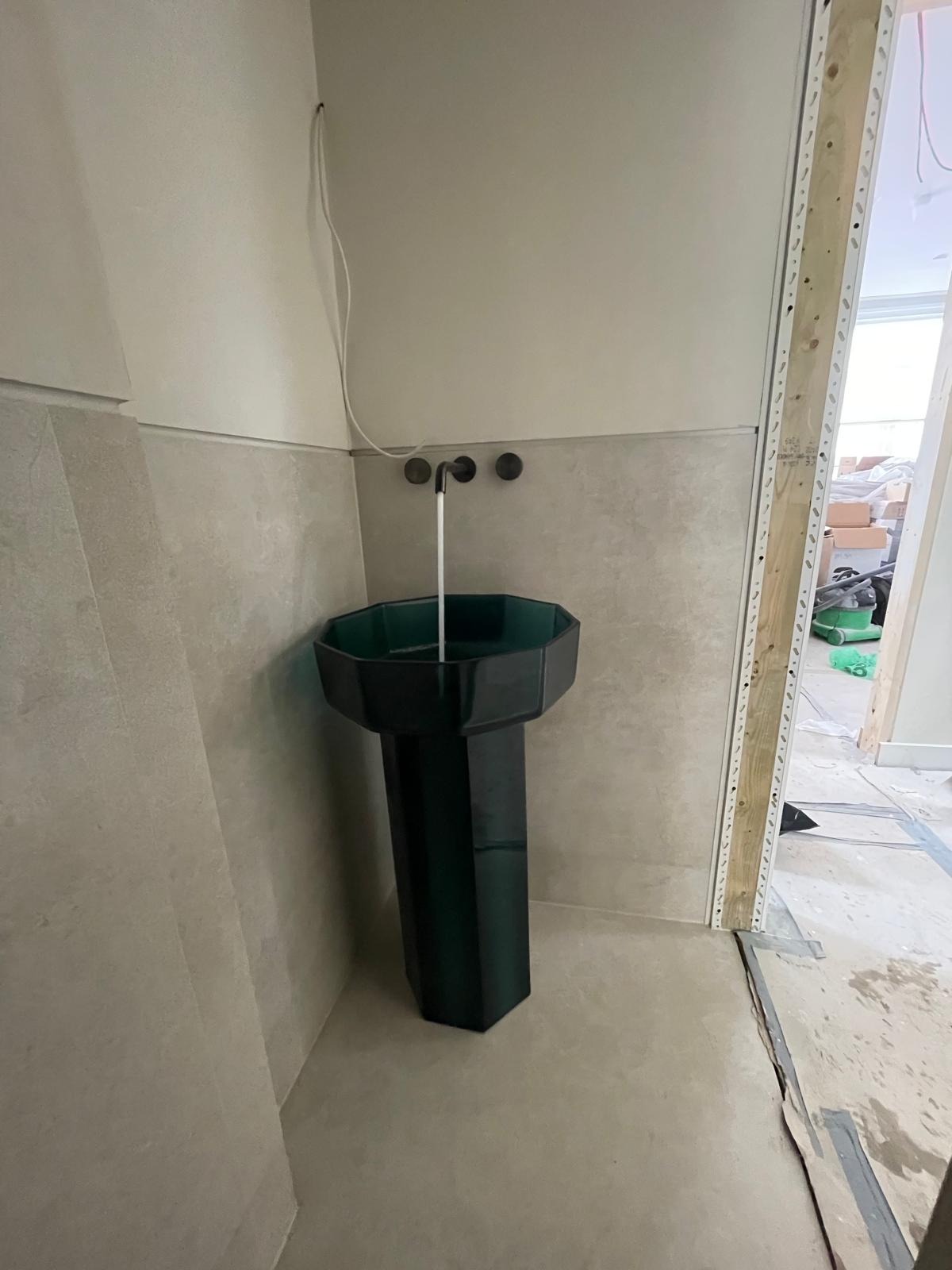 Montaż ciemnozielonej, ośmiokątnej umywalki kolumnowej z trzema czarnymi kranami w łazience w trakcie remontu, widoczne płytki na ścianach, kabel elektryczny i surowe wykończenie ościeżnicy.