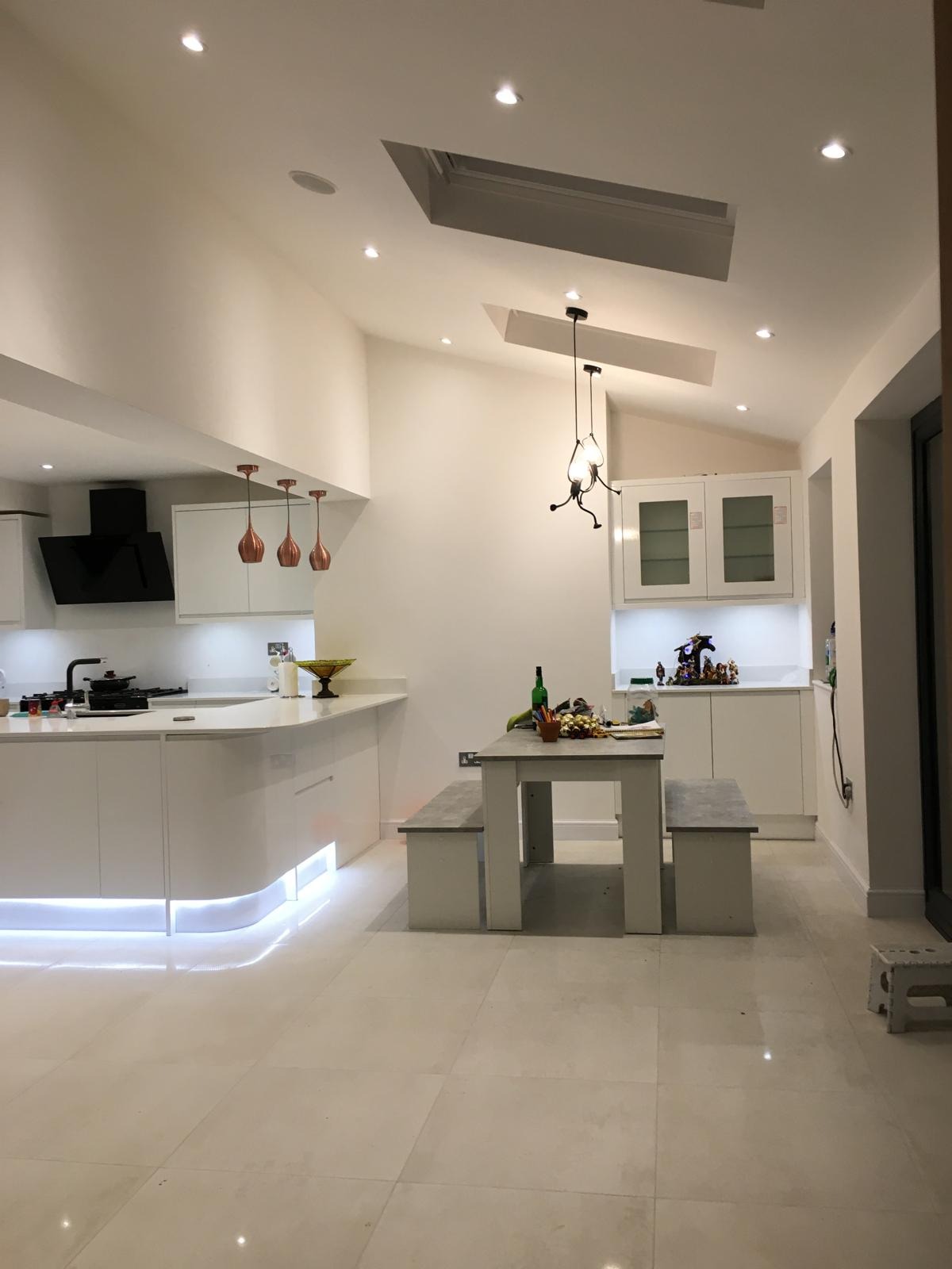 Jasna kuchnia z białymi szafkami, podświetleniem LED pod blatem i stołem z ławkami. Na stole butelka i drobne przedmioty, a na blacie kuchennym owoce w ozdobnej misie. Sufit z oknami dachowymi...