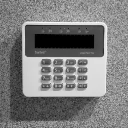 Centrala alarmowa Satel Perfecta, zamontowana na teksturowanej ścianie, widok z przodu, czarno-białe zdjęcie.