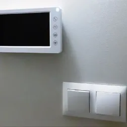 Biały wideofon z czarnym ekranem zamontowany na jasnej ścianie obok podwójnego włącznika światła.