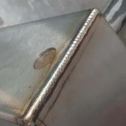 Szczegółowe zbliżenie na spoinę na nierdzewnej konstrukcji, widoczna moneta obok spoiny dla skali.