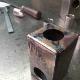 Michal Firlewicz - Metalowy prostopadłościan z otworami, z widocznymi śladami spawania i opalizującymi przebarwieniami, leżący na metalowym stole warsztatowym obok narzędzi ślusarskich.