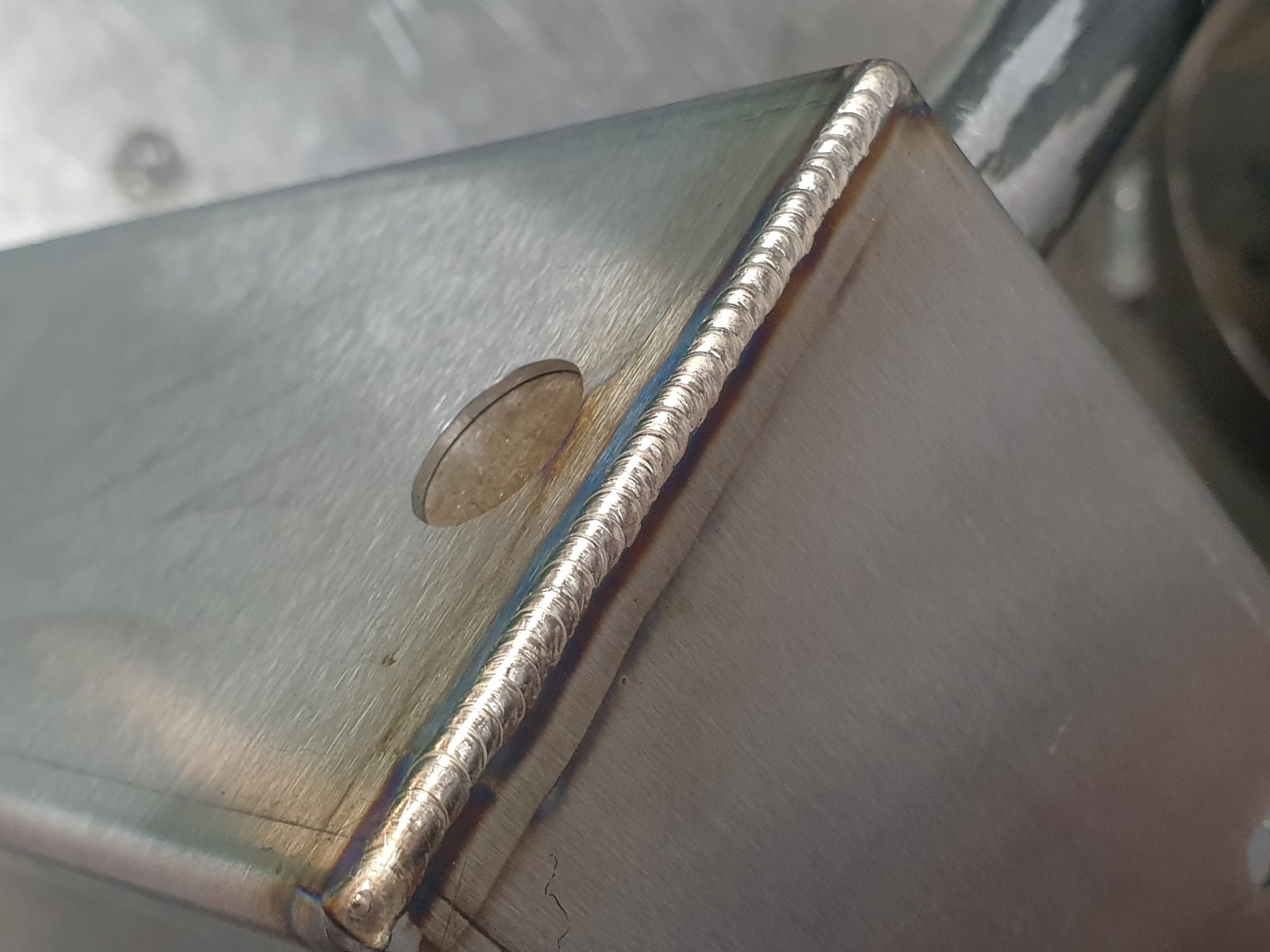 Szczegółowe zbliżenie na spoinę na nierdzewnej konstrukcji, widoczna moneta obok spoiny dla skali.